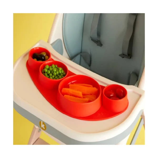 Red silicone placemat with integrated bowls containing sliced carrots, peas, olives, and dipping sauce on a high chair tray.