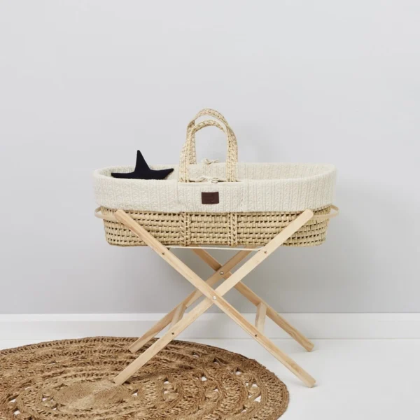 Woven baby bassinet with knitted lining on a foldable wooden stand, placed on a circular woven rug against a white wall.