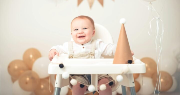 Baby Milestones: A smiling baby in light-coloured overalls sits in a high chair decorated with a garland of pom-poms, next to a paper party hat. The background features gold and white balloons, with a triangular banner hanging above.