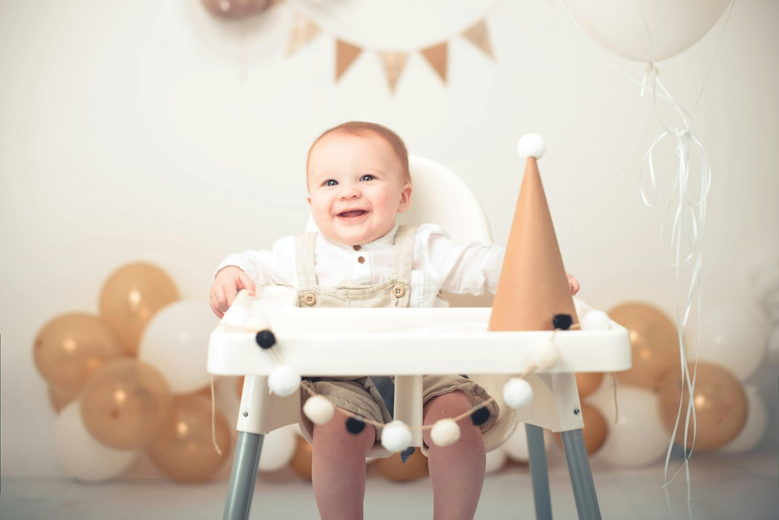 Baby Milestones: A smiling baby in light-coloured overalls sits in a high chair decorated with a garland of pom-poms, next to a paper party hat. The background features gold and white balloons, with a triangular banner hanging above.