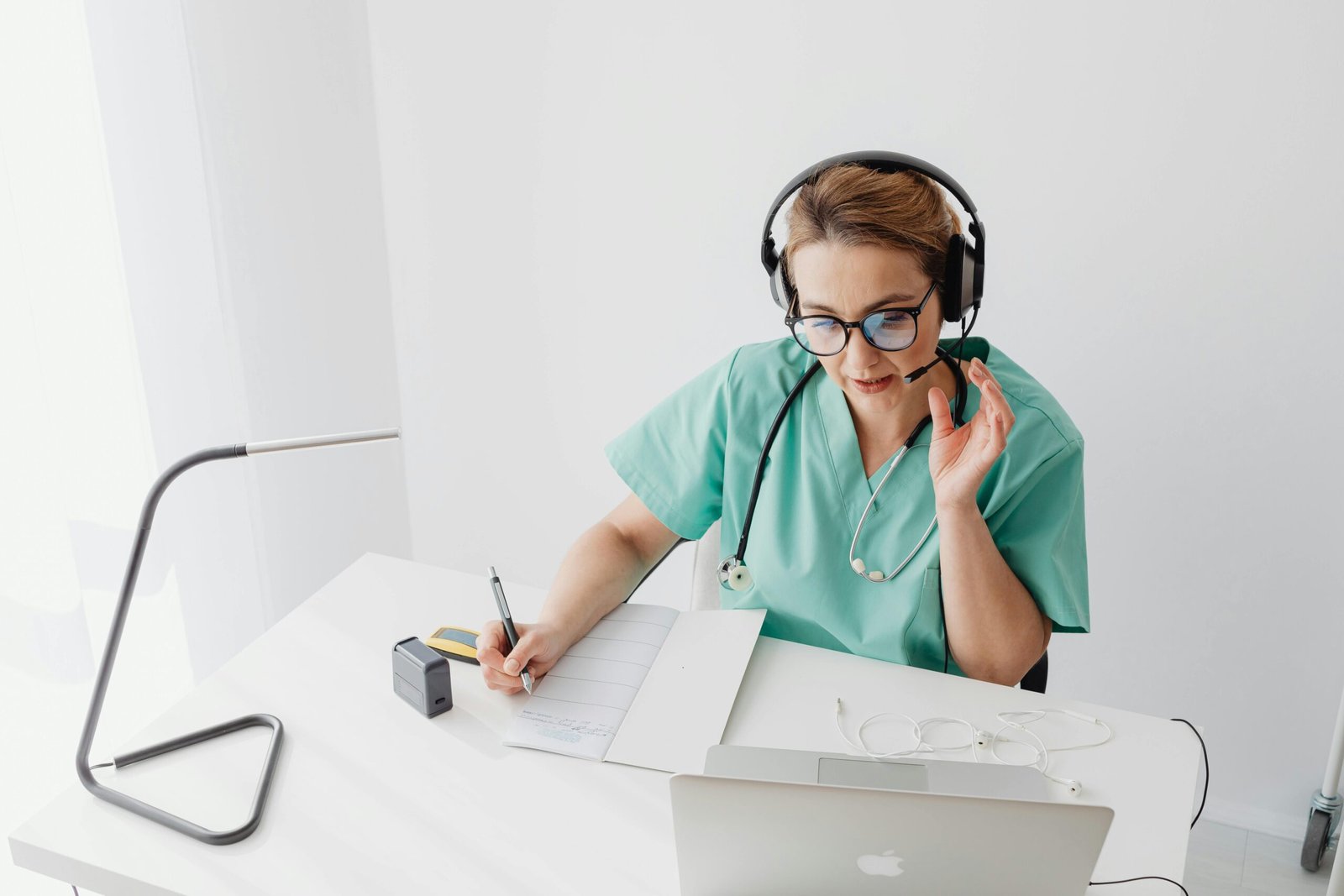 A healthcare professional wearing scrubs and a stethoscope is sitting at a desk, using a laptop and a headset, while taking notes on a clipboard.