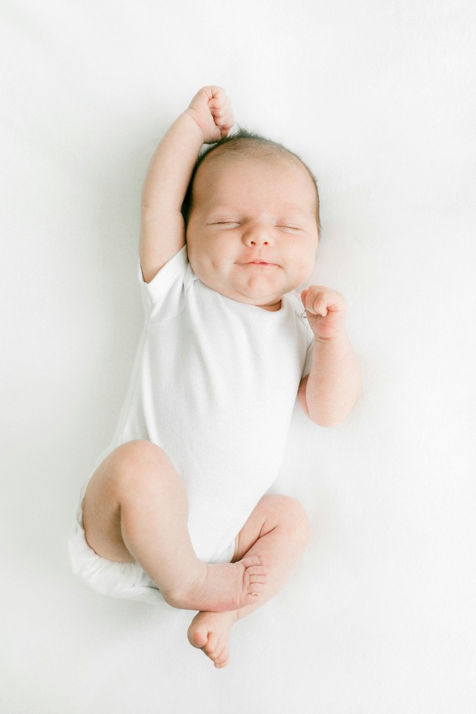 A sleeping baby in a white onesie lies on a white surface, with one arm stretched above the head and the other bent near the face.