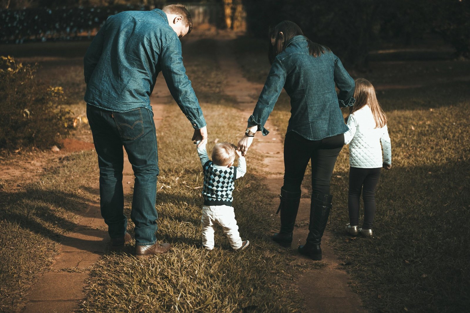 Sibling Room Sharing v Separate Bedrooms 2 Sibling Room Sharing v Separate Bedrooms: A family of four walking on a grassy path, with two adults holding the hands of a toddler in the middle, while a young girl walks alongside.