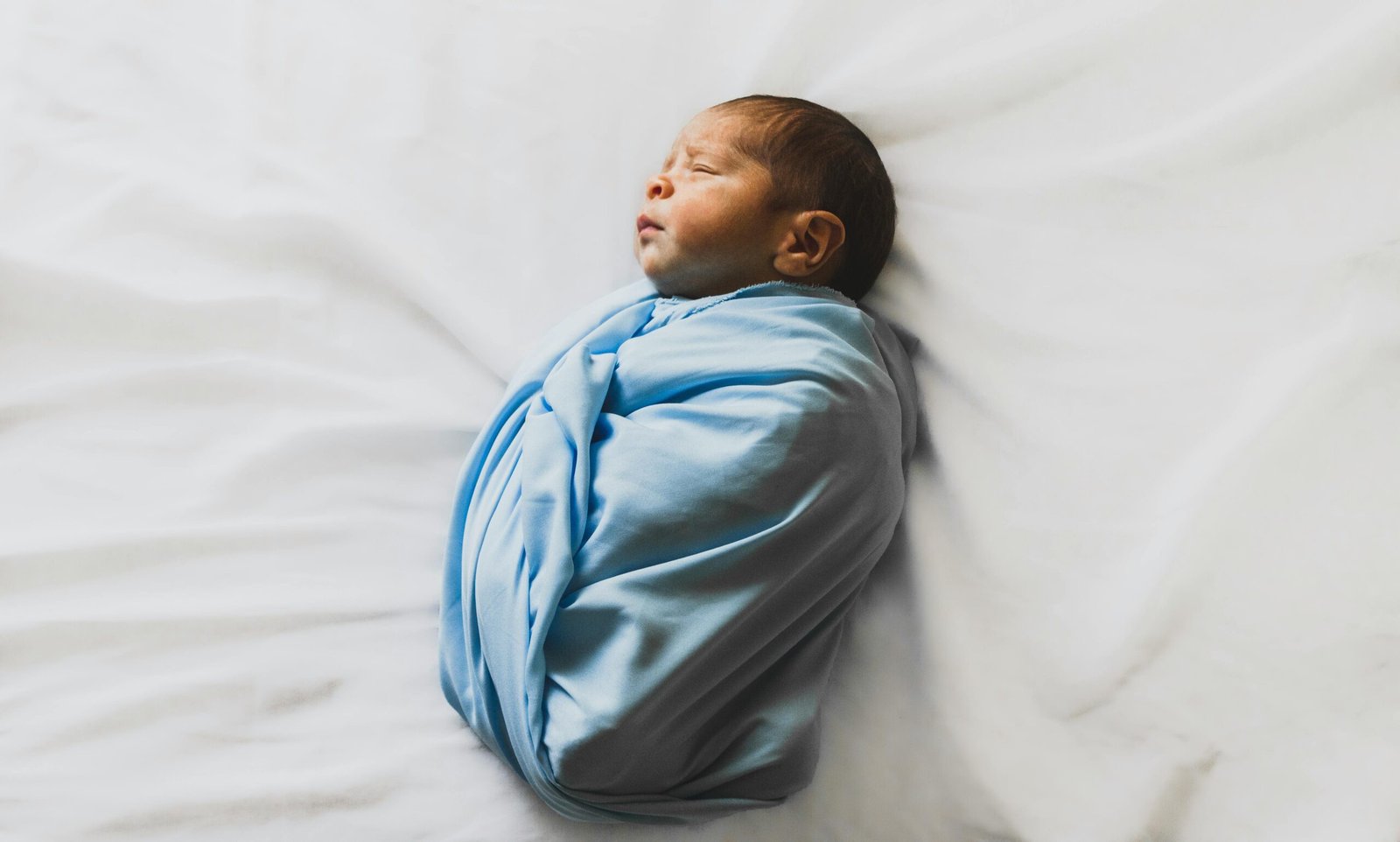 The Ultimate New Baby Hospital Bag Checklist 2 A newborn baby wrapped in a light blue blanket, peacefully asleep on a white bed.