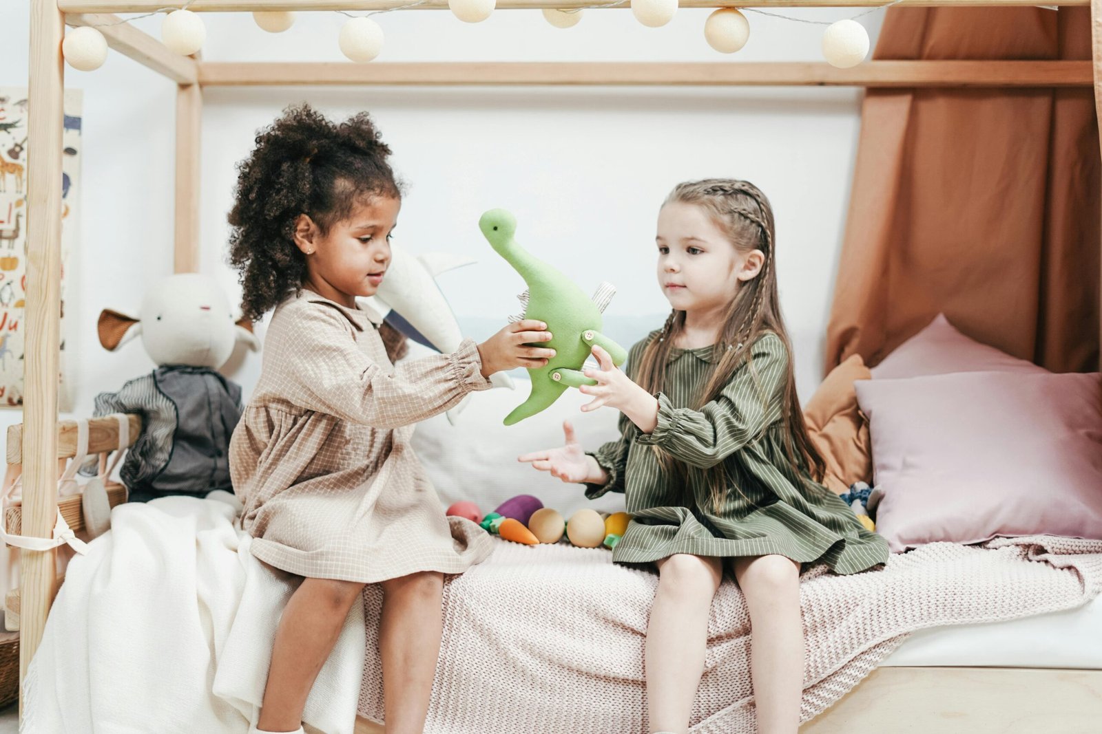 Two young girls sitting on a bed, playing with a green dinosaur plush toy in a cozy room with soft lighting and pillows.