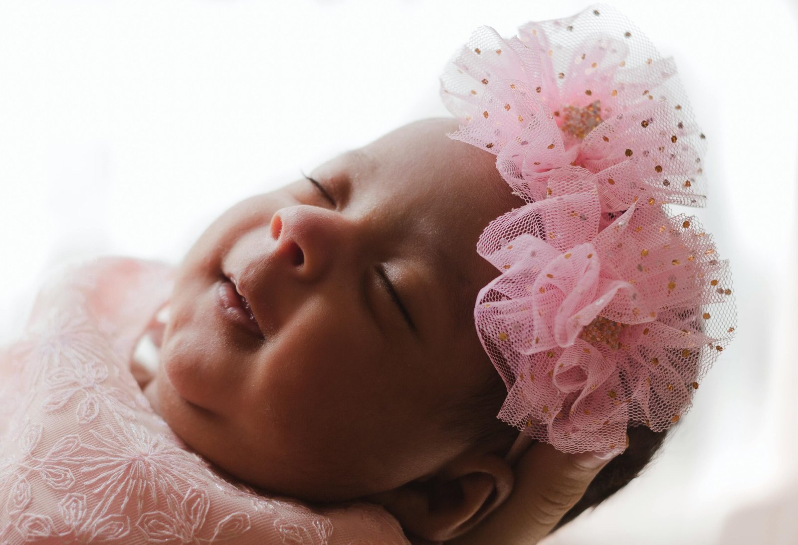 Understanding Baby Sleep Cycles: A sleeping baby with a pink floral headband, gently cradled in a hand, wearing a pink embroidered outfit.